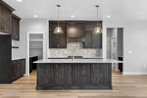 Kitchen featuring dark brown cabinets, light stone counters, tasteful backsplash, a center island with sink, and pendant lighting