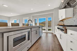 Kitchen with stainless steel appliances, white cabinets, wall chimney range hood, backsplash, and light wood finished floors