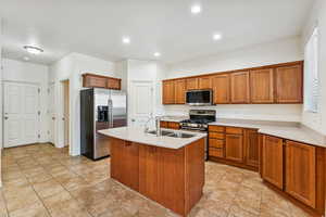 Kitchen featuring stainless steel appliances, brown cabinetry, light stone countertops, light tile patterned flooring, and a center island with sink