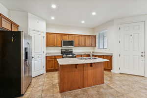 Kitchen featuring brown cabinetry, stainless steel appliances, recessed lighting, light tile patterned floors, and a kitchen island with sink