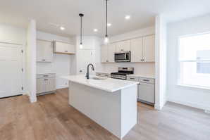 Kitchen with stainless steel appliances, an island with sink, light wood-style flooring, hanging light fixtures, and light stone countertops