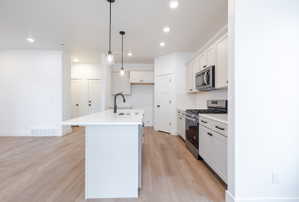 Kitchen with stainless steel appliances, light wood-style floors, an island with sink, backsplash, and white cabinetry