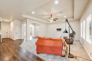 Living room with a raised ceiling, arched walkways, wood finished floors, a ceiling fan, and recessed lighting