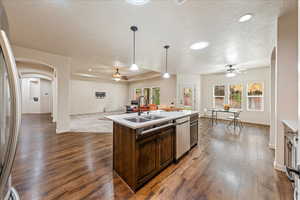Kitchen featuring arched walkways, dark brown cabinets, ceiling fan, an island with sink, and hanging light fixtures