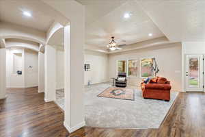 Living room featuring arched walkways, a raised ceiling, dark wood-style floors, a ceiling fan, and recessed lighting