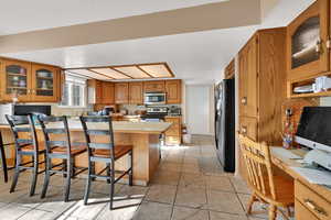 Kitchen featuring brown cabinetry, a kitchen bar, glass insert cabinets, light countertops, and appliances with stainless steel finishes