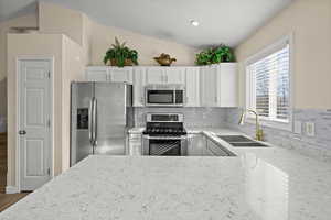 Kitchen with light stone countertops, stainless steel appliances, decorative backsplash, white cabinetry, and lofted ceiling