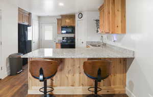 Kitchen featuring a breakfast bar, light stone counters, black appliances, a peninsula, and dark wood-style flooring