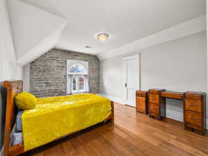 Bedroom with wood finished floors, vaulted ceiling, and brick wall