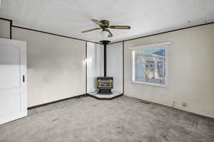 Unfurnished living room featuring a wood stove, carpet floors, a ceiling fan, and ornamental molding