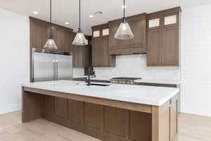 Kitchen with appliances with stainless steel finishes, a center island with sink, dark brown cabinetry, tasteful backsplash, and light stone countertops