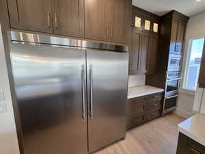 Kitchen with dark brown cabinetry, built in appliances, glass insert cabinets, and light wood finished floors