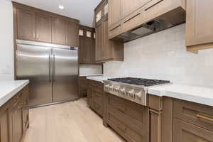 Kitchen featuring ventilation hood, stainless steel appliances, light wood-style floors, decorative backsplash, and dark brown cabinetry