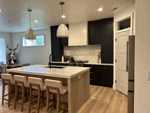 Kitchen featuring dark cabinets, backsplash, decorative light fixtures, a kitchen island with sink, and light stone countertops