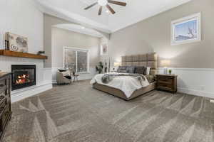 Carpeted bedroom featuring a wainscoted wall, crown molding, a large fireplace, multiple windows, and ceiling fan