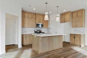 Kitchen featuring wood cabinets to the ceiling, tile backsplash, pendant lighting, appliances with stainless steel finishes,  wood-style flooring, and a center island with sink and upgraded quartz tops