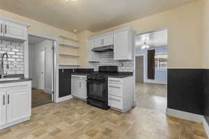 Kitchen featuring black gas range oven, white cabinets, backsplash, under cabinet range hood, and open shelves