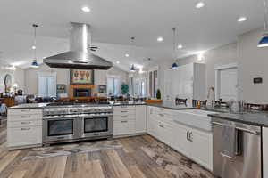 Kitchen featuring stainless steel appliances, range hood, dark stone countertops, hanging light fixtures, and open floor plan