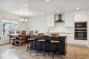 Kitchen featuring decorative backsplash, a breakfast bar, stainless steel oven, wall chimney range hood, and white cabinetry