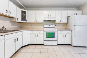 Kitchen featuring white appliances, light countertops, glass insert cabinets, under cabinet range hood, and light tile patterned floors