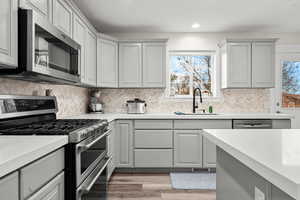 Kitchen featuring stainless steel appliances, gray cabinets, tasteful backsplash, light wood-style flooring, and light stone countertops