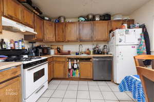 Kitchen featuring white appliances, light tile patterned flooring, under cabinet range hood, brown cabinets, and light countertops