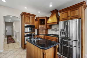 Kitchen featuring stainless steel appliances, dark stone counters, custom range hood, recessed lighting, and an island with sink