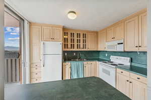 Kitchen with dark countertops, white appliances, glass insert cabinets, light brown cabinetry, and a textured ceiling