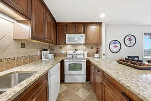 Kitchen with white appliances, backsplash, light stone counters, recessed lighting, and brown cabinetry