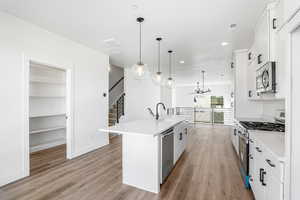 Kitchen featuring stainless steel appliances, white cabinets, a center island with sink, decorative light fixtures, and light wood-type flooring