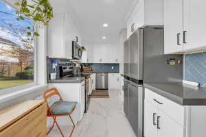 Kitchen featuring dark countertops, backsplash, stainless steel appliances, light marble finish flooring, and white cabinetry