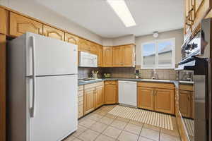 Kitchen with white appliances, light tile patterned floors, backsplash, light brown cabinetry, and dark countertops