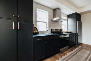 Kitchen featuring dark cabinetry, stainless steel appliances, wall chimney range hood, and dark wood-style floors