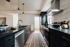 Kitchen featuring dark cabinetry, wall chimney range hood, appliances with stainless steel finishes, and decorative light fixtures