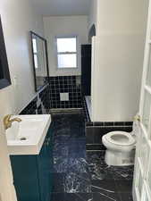 Full bath with tile walls, vanity, wainscoting, and dark marble finish flooring