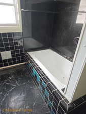 Full bath with a relaxing tiled tub, dark marble finish flooring, and tile walls