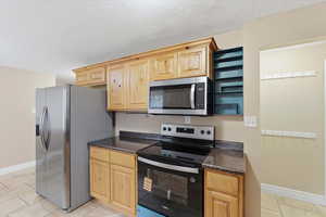 Kitchen with appliances with stainless steel finishes, light tile patterned floors, light brown cabinetry, dark stone counters, and a textured ceiling