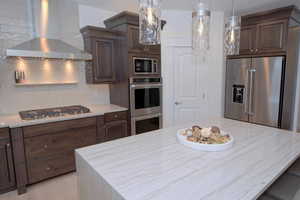 Kitchen featuring wall chimney range hood, appliances with stainless steel finishes, a center island, decorative backsplash, and light stone counters