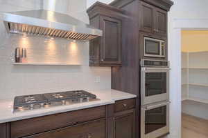 Kitchen with wall chimney exhaust hood, stainless steel appliances, dark brown cabinetry, and light wood-style floors