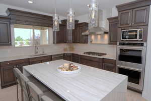 Kitchen with appliances with stainless steel finishes, light stone counters, wall chimney range hood, backsplash, and dark brown cabinets