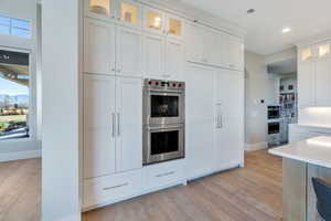 Kitchen with glass insert cabinets, stainless steel double oven, white cabinetry, light wood finished floors, and light stone countertops