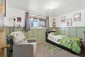 Bedroom with a decorative wall, a textured ceiling, light carpet, and a wainscoted wall