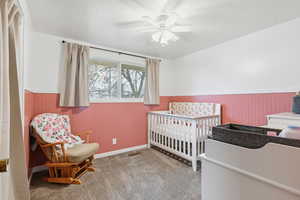 Carpeted bedroom featuring wainscoting, a crib, a textured ceiling, a ceiling fan, and wooden walls