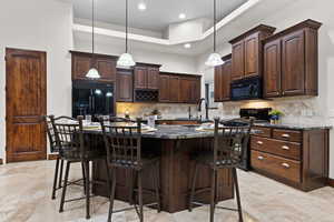 Kitchen with dark brown cabinetry, dark stone counters, hanging light fixtures, black appliances, and an island with sink