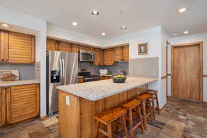 Kitchen featuring brown cabinets, appliances with stainless steel finishes, a kitchen bar, tasteful backsplash, and light stone countertops