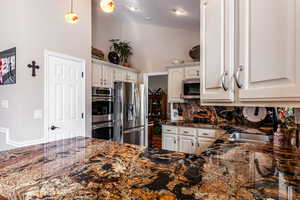 Kitchen featuring dark stone counters, decorative backsplash, stainless steel appliances, hanging light fixtures, and white cabinets