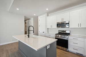 Kitchen with gray cabinetry, stainless steel appliances, a kitchen island with sink, light wood-type flooring, and light stone countertops
