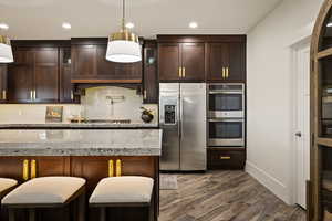 Kitchen with dark brown cabinetry, stainless steel appliances, light stone counters, decorative backsplash, and pendant lighting