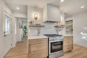Kitchen with stainless steel gas range, open shelves, custom range hood, light brown cabinetry, and light stone countertops