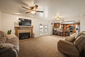 Living area featuring a textured ceiling, a tiled fireplace, light colored carpet, crown molding, and a ceiling fan
